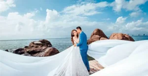 A couple embracing on a rocky shoreline with flowing fabric captured by a Vietnam wedding photographer, showcasing the dramatic coastal beauty of Nha Trang.