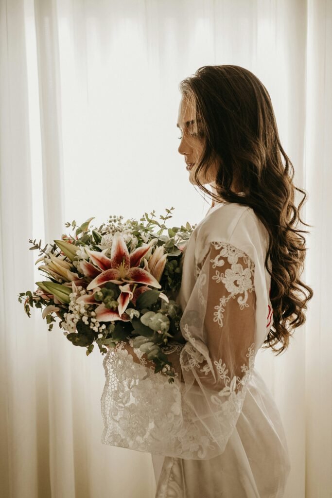 Bride holding a bouquet in soft natural light wearing a lace robe.