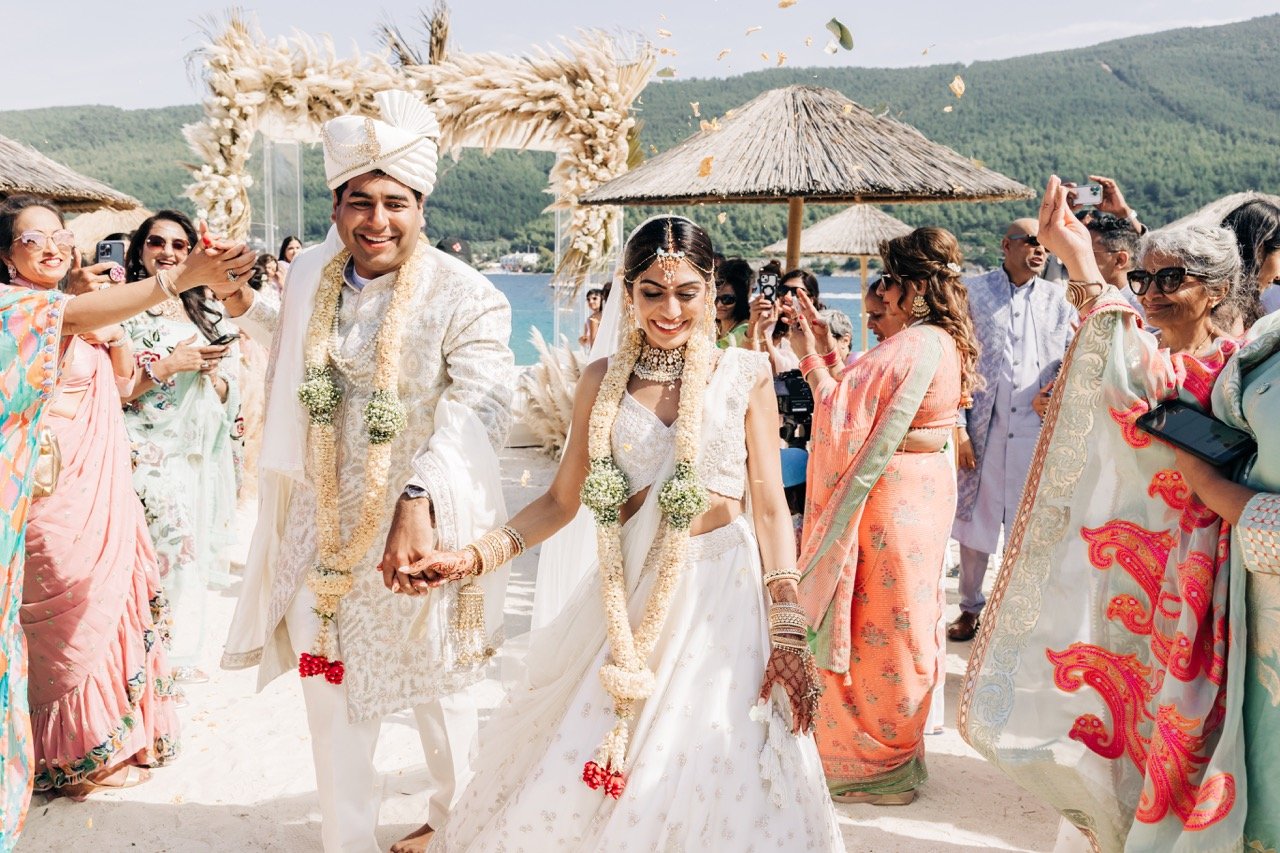 Joyful Indian couple walking hand in hand after their beach wedding ceremony in the South of France, surrounded by guests celebrating under the sun — beautifully captured by a Destination Wedding Photographer in France.